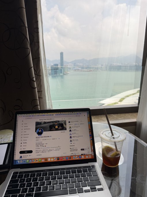 A MacBook Pro laptop open on a table beside a glass of iced tea, displaying an X/Twitter page, with a scenic view of a harbor, city skyline, and mountains through the window.