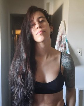 A selfie of a woman with long dark hair wearing a black bikini top, standing in a room with a tattoo visible on her upper arm.