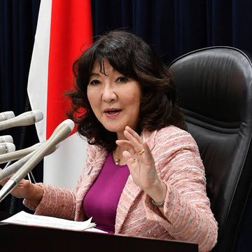 Japanese politician Seiko Noda speaks at a podium with multiple microphones, gesturing with her hand while wearing a pink textured blazer, with a Japanese flag visible in the background.