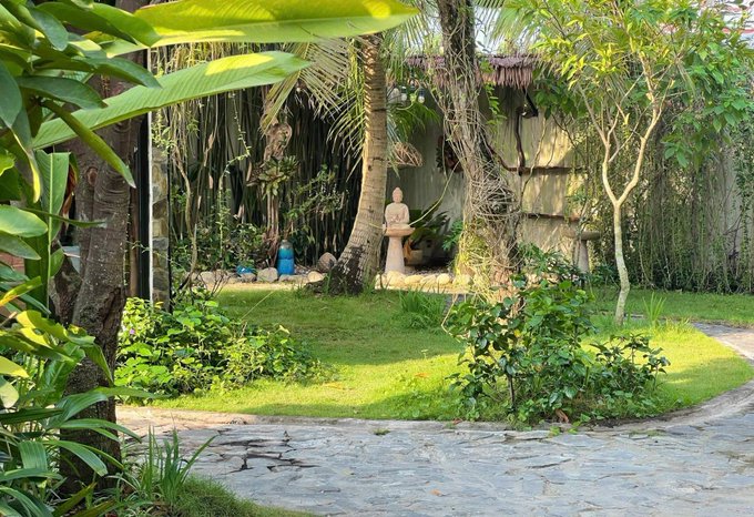 A serene tropical garden with lush green grass, palm trees, and a small statue in the background, with a stone path winding through.