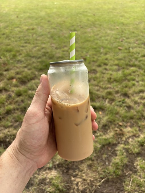 A hand holding a clear can filled with iced coffee (or milk tea) and a green-and-white striped straw, set against a blurred grassy outdoor background.