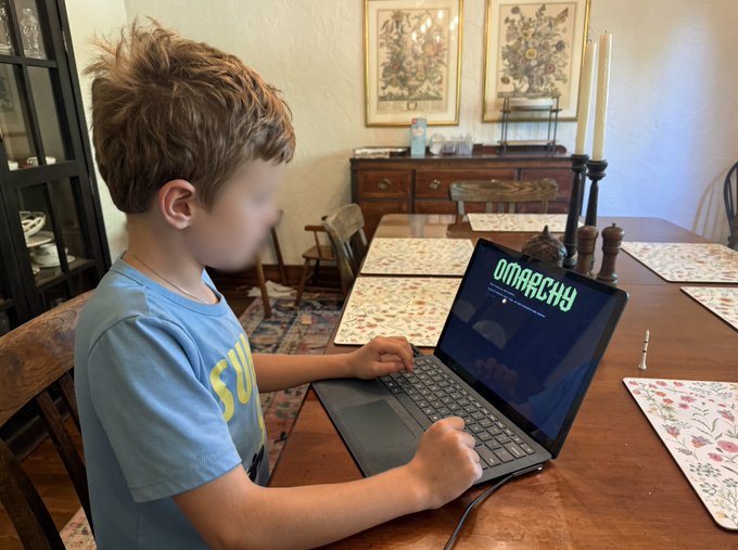 A young boy sits at a wooden dining table using a tablet displaying a website titled 'OMARCHY'.