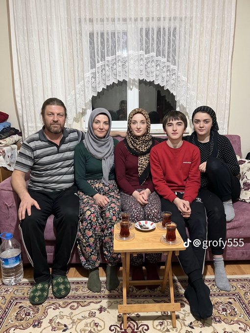 A family of five sits together on a pink couch in a living room, drinking tea from traditional Turkish tulip glasses on a small wooden table, with a TikTok watermark reading "@grofy555".