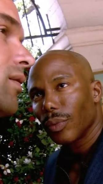 The image shows two men, one of whom has a bald head and the other with a mustache. They are both looking at something off-camera, possibly engaged in conversation or observing an interesting object. The background features a window that allows natural light into the room, creating a warm atmosphere.