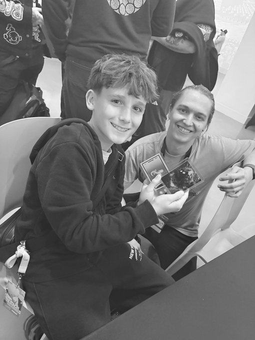A black and white photo of a smiling young boy with braces sitting next to another person, holding what appears to be a Pokémon or trading card.