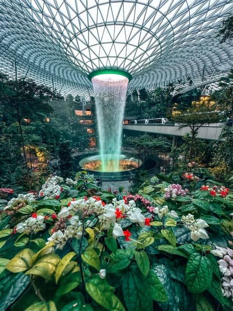 An indoor waterfall pouring from a circular opening in a domed glass ceiling into a lush garden filled with greenery and flowers, likely at Jewel Changi Airport.