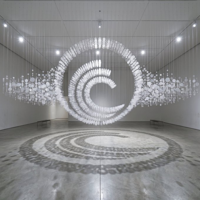 An immersive art installation featuring a suspended spiral 'C' made of white circular discs, casting a matching spiral shadow on the gallery floor.