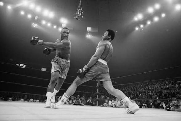 A black-and-white image of a boxing match between two fighters under bright stadium lights, with a crowd visible in the background.
