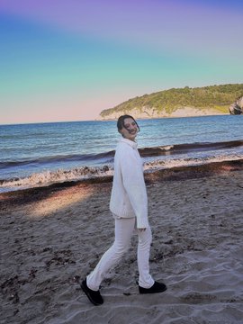 A man dressed entirely in white clothing (sweater and pants) standing on a sandy beach with the ocean and a distant cliff/headland visible in the background under a blue sky.
