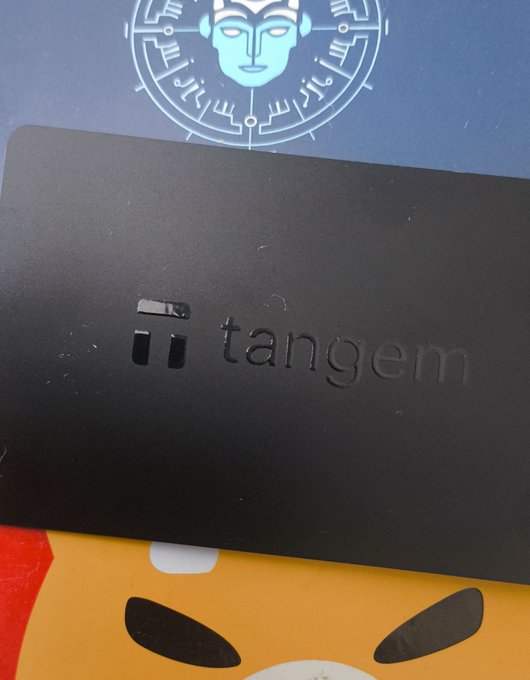 A black metal card embossed with the word "tangem" rests against a blue box featuring a stylized line-art face design and a yellow character with angry eyes.