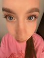 A close-up selfie of a woman with large eyes wearing a pink top.