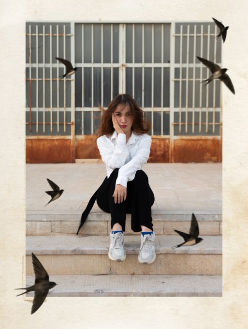 A young woman with shoulder-length hair sits on stone steps in a white shirt and black pants, with swallows digitally added flying around her.