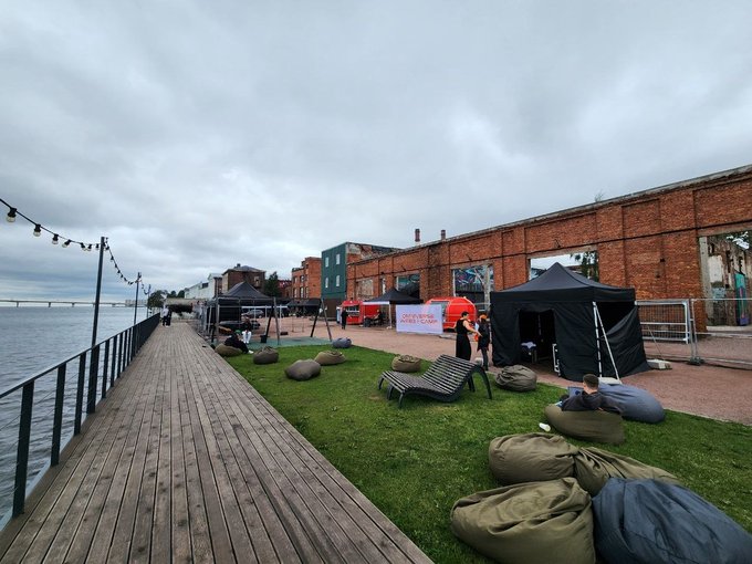 A wooden waterfront boardwalk stretches alongside a sparse outdoor event setup, with only a handful of attendees scattered near tents and bean bags against the backdrop of an old brick industrial building under a grey sky—visually reinforcing the post's lament about dwindling conference attendance and the once-vibrant industry now reduced to mostly empty halls. A partially visible banner reads "IT..." and the black tent is labeled "AVA," while a red inflatable arch and metal barricades further emphasize the underwhelming turnout.