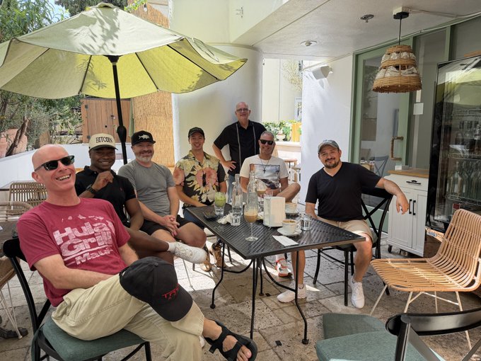 A group of seven men gather around a sunlit patio table beneath a yellow umbrella, enjoying drinks and conversation together. The man in the foreground sports a red "HULA GIRLS Maui" shirt, while another wears a "BITCOIN" cap—fitting the post's theme of finding happiness at a local Bitcoin meetup.