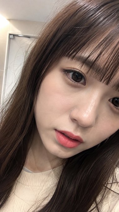 A close-up selfie of a young woman with long brown hair, bangs, and glossy pink lipstick.