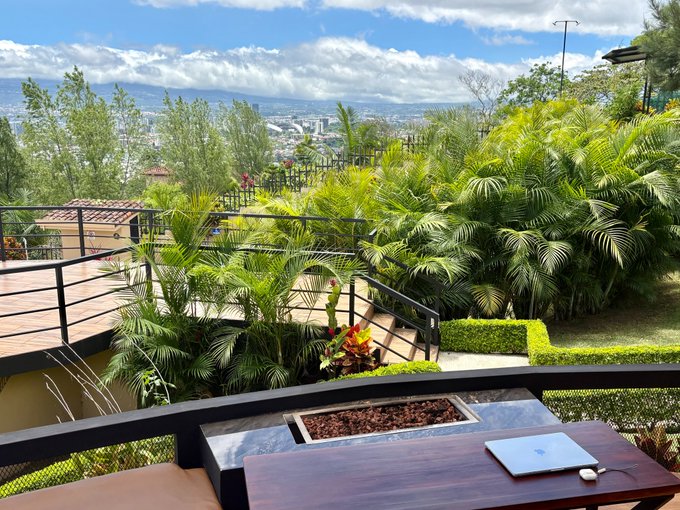 An outdoor patio with a fire pit, wooden table, and MacBook overlooks a tropical landscape with palm trees, manicured gardens, and a distant city under a partly cloudy sky.