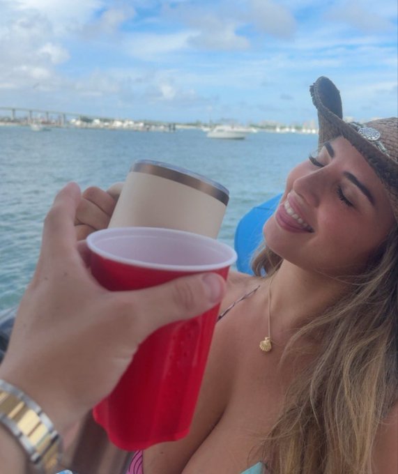 A woman in a cowboy hat and bikini smiles while clinking drinks—a white tumbler and a red solo cup—on a boat with a coastal backdrop. The carefree, tropical vibe of the image clashes humorously with the caption's ironic mix of beach fun and financial markets talk.