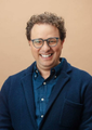 The image features a man wearing glasses, a blue jacket, and a blue shirt with white buttons. He has curly hair and is smiling at the camera. The background of the image is a light beige color that contrasts with his dark clothing. There are no other objects or people in the image, making him the sole focus. The caption "Everyone be ready." suggests that there might be an event or gathering happening soon, possibly related to the post about Mythos dropping tomorrow.