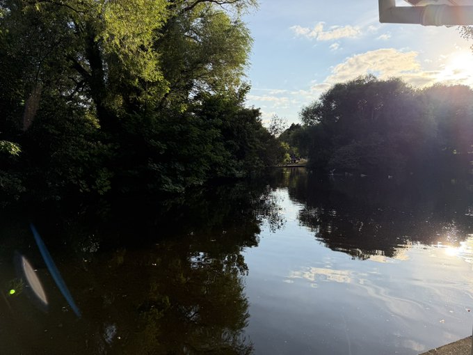 A serene river flows through dense, tree-lined banks under a partly cloudy sky, with the sun creating a bright glare on the right side and casting reflections across the calm water. A white pipe is visible in the upper right corner, and a small dark object protrudes from the water on the lower left.