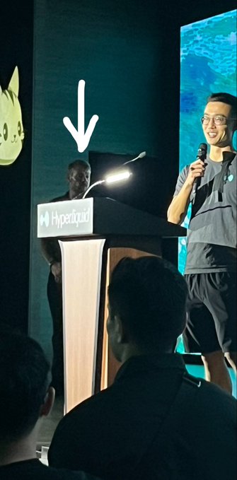 A man with white hair stands behind a podium labeled "Hyperliquid" on a stage, while another speaker addresses an audience with a microphone, with an arrow in the image pointing to the man in the background—likely the Michael Saylor referenced in the post about the $320M $HYPE buyer.