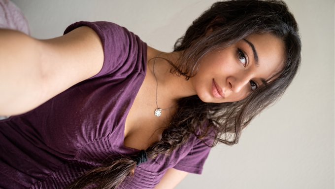 A young woman with long dark braided hair and a delicate pendant necklace poses for a selfie while wearing a purple v-neck top.