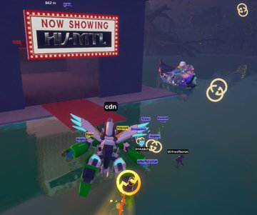 A screenshot from a 3D video game or virtual world showing a mech robot character in front of a "NOW SHOWING" theater sign, with other players visible in the scene.