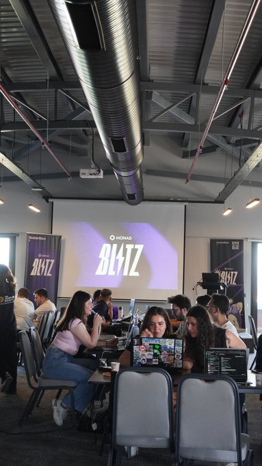Hackers gather around laptops at a coding event featuring "BLITZ" and "MONAD" branding on large screens in an industrial-style venue. The scene captures the Turkish developer community actively building with Monad at the Blitz Ankara hackathon.