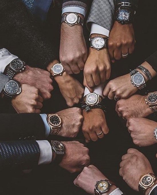 A circle of hands wearing various luxury watches meeting in the center, showcasing different timepieces on suited wrists.