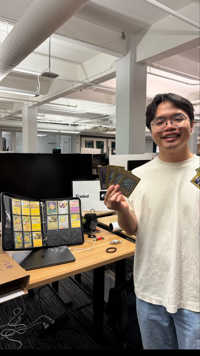 A smiling man in glasses stands in an office holding up several Pokemon cards, with a binder full of yellow Pikachu-themed cards visible on the desk beside a black monitor.