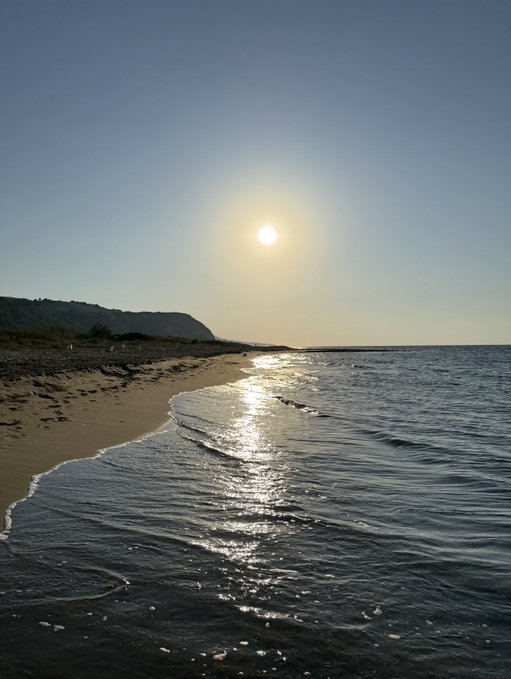 A serene beach scene at sunset or sunrise, featuring a bright sun in a clear sky reflecting brilliantly on the calm ocean waves rolling onto a sandy shore, with a dark silhouette of a hill or cliff in the distance.