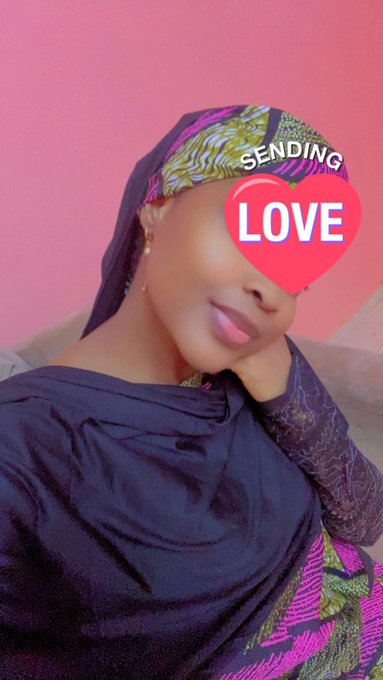 A woman wearing a black hijab and patterned headscarf smiles at the camera with a "SENDING LOVE" heart emoji overlay on her face, against a pink background.
