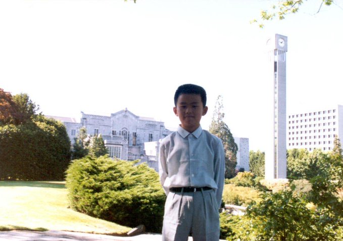 A young boy in a white shirt and gray pants stands posed outdoors on a sunny day, with a tall white clock tower and a stately building visible in the background. The vintage photograph likely represents the author's childhood, symbolically referencing following CZ's (Changpeng Zhao's) footsteps from an early age as a long-time admirer celebrating Binance's 9th anniversary.