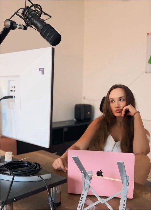 A young woman with long brown hair sitting at a desk with a pink laptop and a microphone, looking thoughtfully off-camera in what appears to be a podcast or streaming setup.