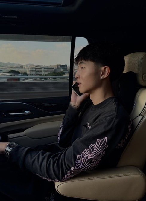 A young man sits in the back seat of a car, wearing a dark long-sleeved shirt with decorative patterns, holding a phone to his ear while looking out the window at a cityscape.