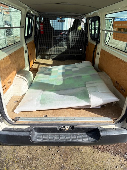 The interior of a converted van showing a plywood floor with white foam padding laid out, presumably for building out a camper van conversion with a sleeping area.
