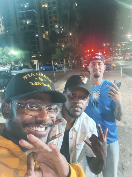 Nighttime selfie of three men posing on a city street, with one wearing a 'THA GREAT' cap and making peace signs.
