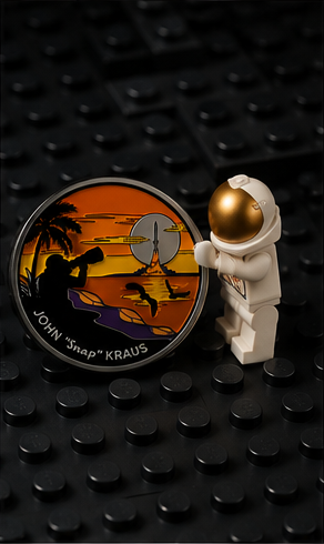 A small LEGO-style astronaut figure with a gold visor stands next to a round pin featuring a surfer sunset scene and the text "JOHN 'snap' KRAUS".