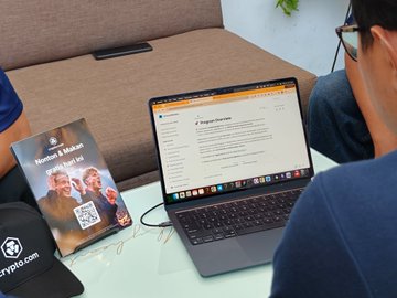 A group gathers around a laptop displaying what appears to be a page from Cronos (cronos-pos.org), with a "Women of Kronos" book nearby, as they discuss the Cronos blockchain following a Crypto.com watch party.