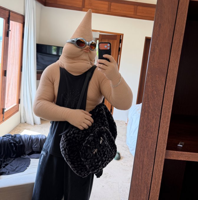 A person wearing a flesh-toned full-body suit that gives the appearance of a phallic shape, complete with novelty glasses and pursed lips, takes a mirror selfie in a bedroom. The image humorously plays on the "Is this $clitcoin?" post by visually merging anatomical references with cryptocurrency slang, creating an absurd meme-style juxtaposition.