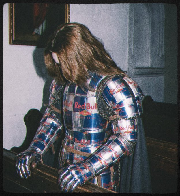 An artistic photo of a person wearing armor made entirely of Red Bull energy drink cans, standing in a dimly lit room next to a framed painting.