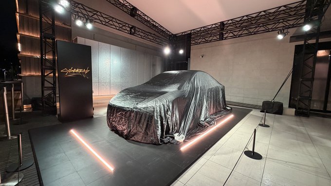 A car is concealed under a dark cover on a display platform illuminated by orange lights, with a 'CYBERCAB' sign visible at an indoor exhibition space.