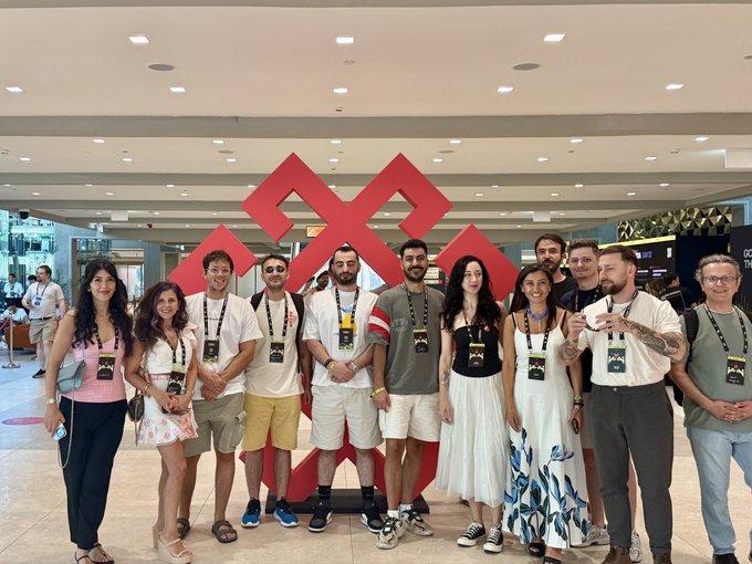 The SuperteamTR crew and ecosystem friends pose together at the Serbia Summit, smiling in front of a large red geometric sculpture in a modern convention center. The group wears matching conference lanyards, reflecting the cross-border collaboration highlighted in the post's appreciation to @SuperteamBLKN.