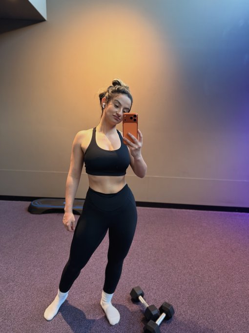 A mirror selfie of a woman in a black sports bra and leggings standing in a gym with purple carpet, holding an orange smartphone, with a pair of dumbbells visible on the floor.