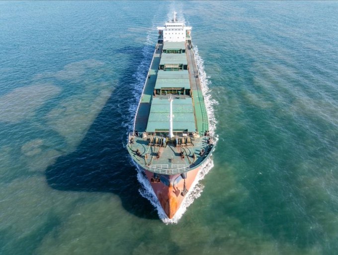 An aerial view of a large cargo ship sailing through clear blue-green ocean waters, casting a long shadow on the sea surface.