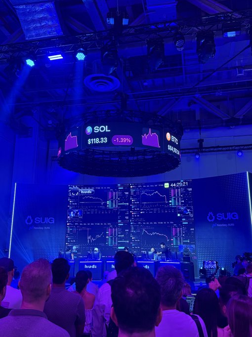 A large crowd attends a crypto conference at the SUIG venue, illuminated by blue lights, with screens displaying 'SOL $118.33 -1.39%' and 'BTC $60,167' alongside trading charts.