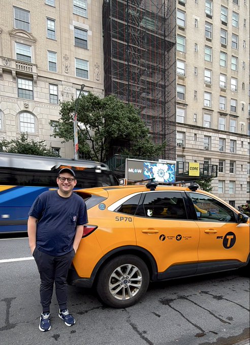A man wearing glasses, a black cap, and a dark t-shirt standing on a New York City street beside a yellow NYC taxi (MTA) with 'SP70' on the side, and a bus passing behind him.