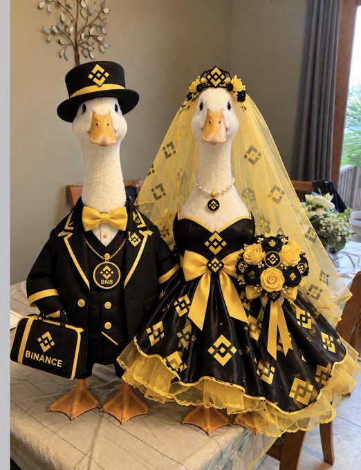 Two goose figurines dressed in wedding attire; the left goose wears a black and yellow suit with a top hat and a briefcase labeled "BINANCE", while the right goose wears a black and yellow dress with a veil and holds a bouquet, both featuring BNB (Binance Coin) logos.