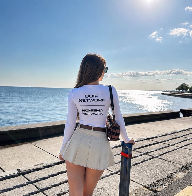 A woman standing by the ocean wearing a white long-sleeve shirt with 'QUIP NETWORK' and 'NOMISMA NETWORK' printed on the back, paired with a white pleated skirt.