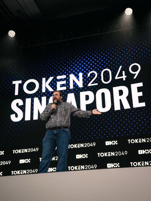 A man with glasses and a beard, wearing a patterned shirt and jeans, stands on stage holding a microphone and gesturing while presenting at the TOKEN 2049 Singapore conference, with the event's branding displayed prominently on the large screen and backdrop behind him.