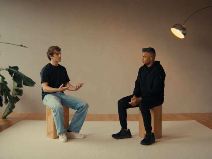 Two men sit on wooden stools facing each other in a minimalist room with a plant, appearing to be in a thoughtful conversation or interview.